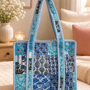 May include: A square quilted tote bag featuring a patchwork design of various blue and white floral and geometric patterns. The bag has two long shoulder straps and edges trimmed with a blue and white striped fabric, creating a cohesive look.