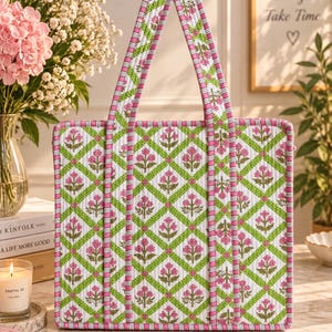 May include: A quilted tote bag featuring a white background with a green diamond grid pattern and pink floral motifs. The bag has two long shoulder straps with pink and white striped trim, which also edges the rectangular body of the bag.