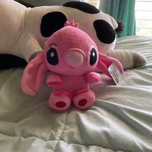 May include: A pink plush toy of the character Angel from Lilo & Stitch, with large black eyes and long ears. The plush is sitting on a light blue bedspread, with a large cow-patterned plush in the background.