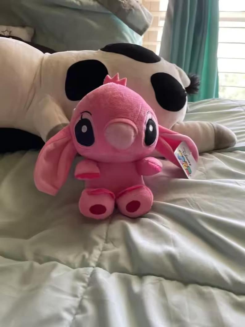 May include: A pink plush toy of the character Angel from Lilo & Stitch, with large black eyes and long ears. The plush is sitting on a light blue bedspread, with a large cow-patterned plush in the background.