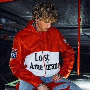 以下が含まれることがあります： 前面に「Lost Americana」とプリントされた赤と白のジャケット。左袖にパッチ、左胸に道路標識パッチが付いています。白いシャツの上にジャケットを着ています。