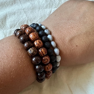 May include: Four bracelets stacked on a wrist. The bracelets are made of different materials, including wood, lava rock, and grey beads.