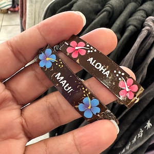 May include: Two brown wooden bracelets with painted flowers and the words "ALOHA" and "MAUI".