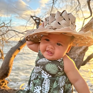 May include: A baby wearing a woven straw hat and a green dress with a white leaf pattern. The baby is smiling, with the ocean and a sunset in the background. The hat is made of woven straw.