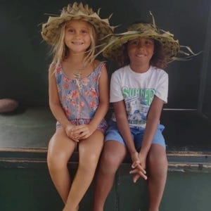 May include: Two children wearing straw hats sit side-by-side, smiling at the camera. One child wears a striped romper with floral accents, the other a white t-shirt with the word "SUMMER" printed on it, and blue shorts. The background is a dark green.