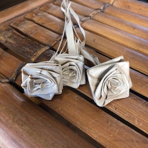 May include: Four beige paper roses tied together with a beige ribbon. The roses are made from woven paper and are arranged in a bouquet.