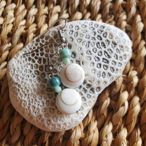 May include: A pair of silver earrings with turquoise and white shell beads. The earrings are hanging from a white coral-like surface.