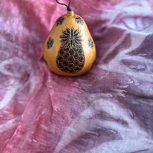 May include: A wooden pear-shaped ornament with a carved pineapple design and black floral accents. The ornament is on a pink and purple fabric background.