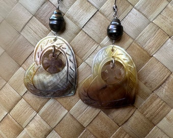 May include: A pair of heart-shaped earrings made from mother of pearl with a carved floral design. The earrings are accented with a black pearl and a silver hook.