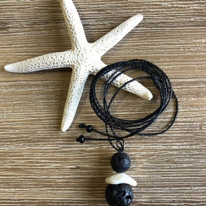 May include: A black lava stone necklace with a white shell charm. The necklace is on a wooden surface with a white starfish.