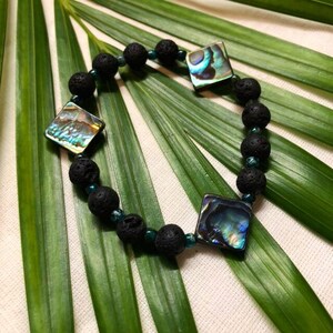 May include: A black lava rock bracelet with three iridescent abalone shell squares. The bracelet is on a green leaf background.