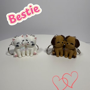 May include: Keychains featuring pairs of cartoon animals: white cats and brown dogs. The cats and dogs are hugging each other. The word "Bestie" is written in pink and white. Two pink hearts are drawn below the keychains.