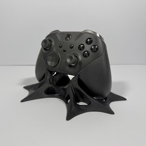 May include: A black Xbox controller rests on a custom-designed black stand. The controller features multiple buttons, joysticks, and textured grips. The stand has a unique, stylized design with a star-like base, providing a secure and stylish display for the gaming accessory.