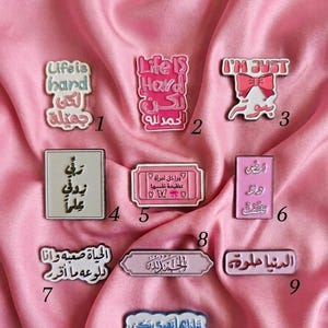 May include: A collection of ten enamel pins featuring various shapes and colours with English and Arabic text. Pin 1 says Life is hard, pin 2 says Life IS Hard, pin 3 says I'M JUST, and pins 4 through 10 feature diverse Arabic phrases and decorative motifs.