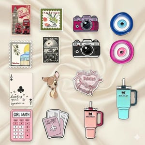 May include: A selection of enamel pins and stickers. The image includes a London postcard, Palestine stamps, cameras, an evil eye, a playing card, a deer, a Pilates heart, a calculator, a passport, and Stanley tumblers.