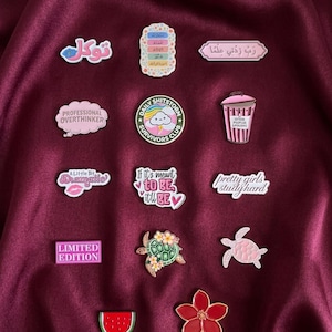 May include: A collection of fourteen enamel pins featuring various designs including Arabic calligraphy, a pink trash can, a turtle with flowers, a watermelon slice, a red flower, and text-based slogans like Professional Overthinker and Limited Edition.