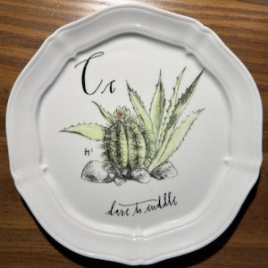 May include: White ceramic plate with a scalloped edge, featuring a hand-drawn cactus illustration in green and yellow. The plate has the word "Ca" and the phrase "dare to cuddle" written on it.