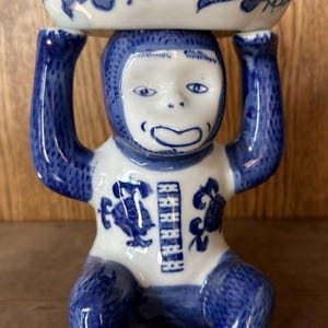 May include: A blue and white ceramic monkey figurine holding a small bowl above its head. The monkey is detailed with blue accents and a white face. The bowl features a floral design.