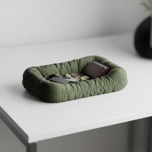 Könnte beinhalten: Eine olivgrüne, rechteckige Ablageschale mit gesteppter Textur. Die Schale enthält ein braunes Portemonnaie, Schlüssel und Münzen. Die Schale steht auf einer weißen Oberfläche.