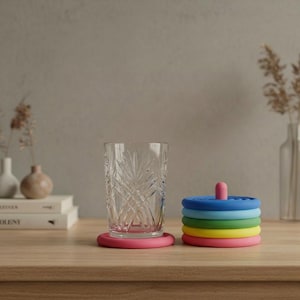 Puede incluir: Un vaso transparente con diseño descansa sobre un posavasos rosa. Al lado, una pila de posavasos coloridos en azul, verde, amarillo y rosa, coronados por una clavija rosa. Tema de decoración del hogar.
