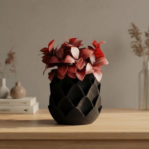 Könnte beinhalten: Eine schwarze, strukturierte dekorative Vase mit einem einzigartigen, geschichteten Design, gefüllt mit leuchtend rotem Laub. Die Vase steht auf einer Holzoberfläche und präsentiert ein modernes, künstlerisches Blumenarrangement. Das Design der Vase ähnelt einer Artischocke.