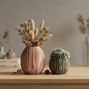 Könnte beinhalten: Zwei dekorative Vasen auf einer Holzoberfläche. Die größere Vase ist pfirsichfarben und enthält Trockenblumen. Die kleinere Vase ist grün und enthält Sukkulenten. Beide Vasen haben ein strukturiertes, geripptes Design.