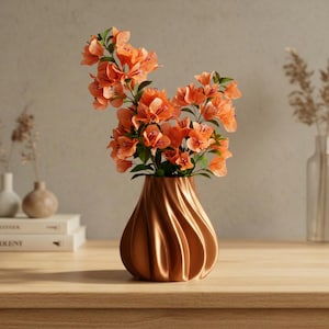 Puede incluir: Un jarrón de color cobre con un diseño en espiral contiene un vibrante arreglo de flores naranjas con hojas verdes. El jarrón está sobre una superficie de madera, creando una estética cálida. Las flores añaden un toque de naturaleza a la decoración.