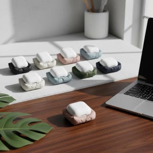 Könnte beinhalten: Eine Sammlung weißer kabelloser Ohrhörer in verschiedenfarbigen, gepolsterten Hüllen. Die Hüllen sind in den Farbtönen Schwarz, Beige, Rosa, Blau, Grün und Marine gehalten. Ein Laptop und ein weißer Keramikbehälter mit Holzutensilien sind im Hintergrund zu sehen.