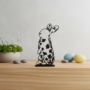 Könnte beinhalten: Schwarze Holz-Osterhasen-Dekoration mit floralem Design. Der Hase steht auf einem kleinen rechteckigen Sockel. Im Hintergrund befinden sich zwei Ostereier, eines blau und eines gelb.