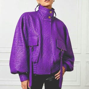 May include: A vibrant purple jacket featuring a textured, woven-look finish, a high collar with two gold-tone buttons, and two large front flap pockets with long, dangling fabric straps. The jacket has voluminous sleeves and a structured silhouette.