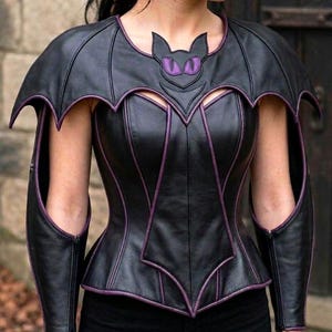 May include: A black corset top featuring purple piping and bat-wing shaped shoulder extensions. The neckline displays a stylized bat face with purple eyes. The garment includes matching arm sleeves with cutouts, all constructed with black panels.