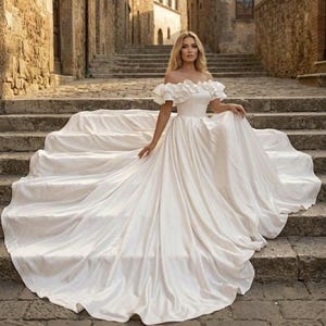 Puede incluir: Un vestido de novia largo hasta el suelo en color blanco marfil con hombros descubiertos. El corpiño presenta un escote con volantes y detalles de tela superpuesta. La falda es voluminosa y amplia, extendiéndose en una larga cola sobre los escalones.