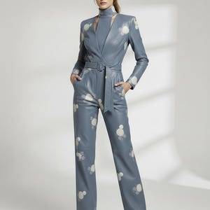 May include: A blue leather jumpsuit with a high neckline and long sleeves. The jumpsuit features a wrap-style top with a belt and wide-leg pants. White floral-like patterns are scattered across the garment. The model is wearing black heels.