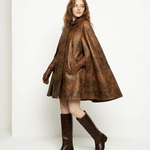 May include: A brown leather cape with a distressed finish, featuring a button-front closure, a small collar, and side arm slits. The garment is paired with matching brown leather gloves and knee-high brown leather boots, creating a cohesive look.