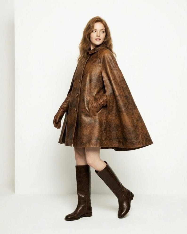 May include: A brown leather cape with a distressed finish, featuring a button-front closure, a small collar, and side arm slits. The garment is paired with matching brown leather gloves and knee-high brown leather boots, creating a cohesive look.