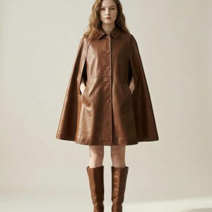 May include: A brown leather cape featuring a classic collar, a front button closure with five visible buttons, and side arm openings. The garment has a structured silhouette that flares slightly toward the hem, paired with matching brown leather knee-high boots.