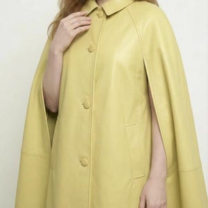 May include: A pale yellow leather cape featuring a classic collar, a front button closure with matching covered buttons, and open arm slits. The garment has a smooth, structured finish and includes vertical welt pockets on the front panels.