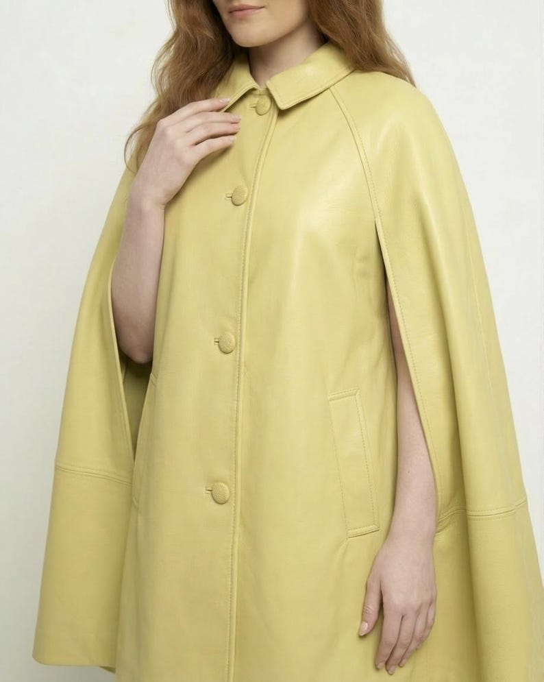May include: A pale yellow leather cape featuring a classic collar, a front button closure with matching covered buttons, and open arm slits. The garment has a smooth, structured finish and includes vertical welt pockets on the front panels.