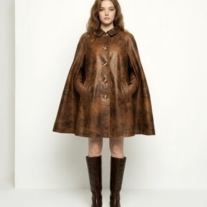May include: A brown leather cape featuring a distressed finish, a classic collar, and a front button closure. The garment has arm slits and two side pockets. It is paired with dark brown knee-high leather boots that reach below the calf.