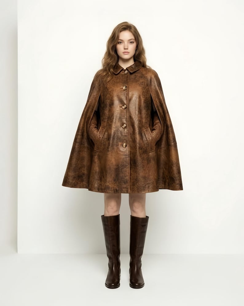 May include: A brown leather cape featuring a distressed finish, a classic collar, and a front button closure. The garment has arm slits and two side pockets. It is paired with dark brown knee-high leather boots that reach below the calf.