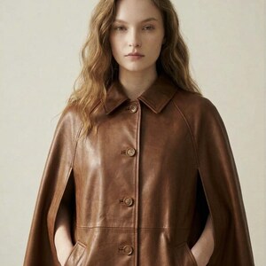 May include: A brown leather cape featuring a classic collar, a front button placket with four visible circular buttons, and side arm openings. The garment has a smooth, polished finish and a structured silhouette that drapes over the shoulders.