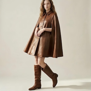 May include: A brown leather cape featuring a classic collar, a front button closure, and arm slits. The garment has a flared silhouette that reaches mid-thigh. It is paired with matching brown leather knee-high boots with a low block heel.