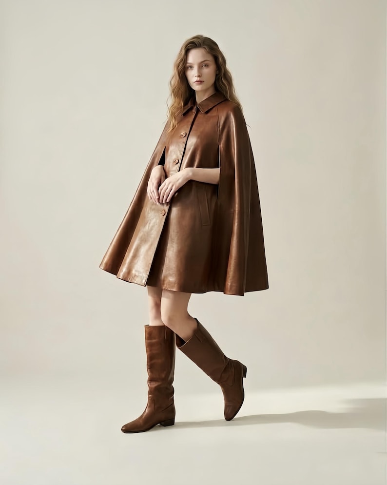 May include: A brown leather cape featuring a classic collar, a front button closure, and arm slits. The garment has a flared silhouette that reaches mid-thigh. It is paired with matching brown leather knee-high boots with a low block heel.