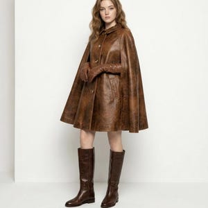 May include: A brown leather cape featuring a collared neckline, a front button closure, and side arm slits. The garment is paired with matching brown leather gloves and knee-high brown leather boots, creating a coordinated ensemble with a textured finish.