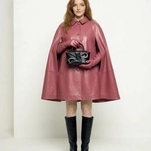 May include: A dusty rose leather cape featuring a classic collar and front button closure, paired with matching leather gloves, black knee-high boots, and a structured black handbag with silver-toned hardware accents on the front panel.