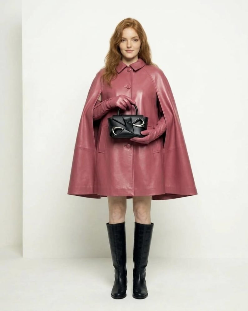 May include: A dusty rose leather cape featuring a classic collar and front button closure, paired with matching leather gloves, black knee-high boots, and a structured black handbag with silver-toned hardware accents on the front panel.