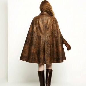 May include: A brown leather cape with a distressed finish, featuring a structured collar, vertical braided seam detailing, and a flared silhouette. The garment is worn with matching brown leather gloves and dark brown knee-high leather boots.