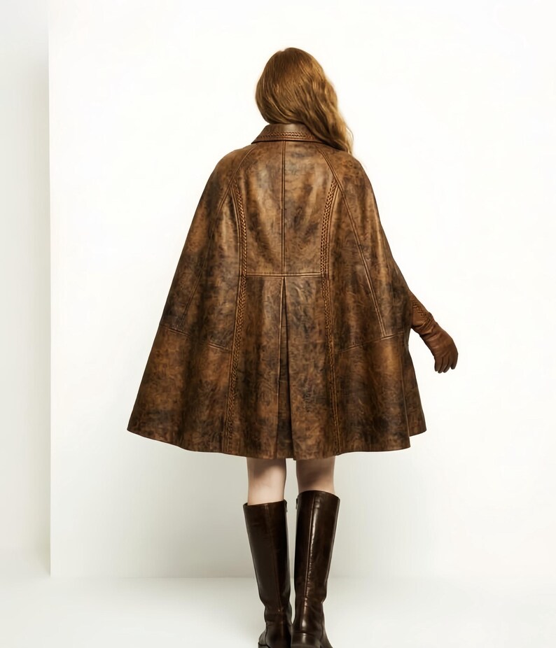 May include: A brown leather cape with a distressed finish, featuring a structured collar, vertical braided seam detailing, and a flared silhouette. The garment is worn with matching brown leather gloves and dark brown knee-high leather boots.