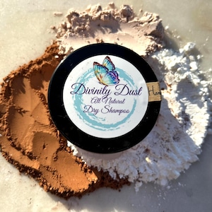 May include: A black container of "Divinity Dust All Natural Dry Shampoo" with a butterfly design, surrounded by brown, tan, and white powder. The container has a small, rectangular label that says "Handmade".