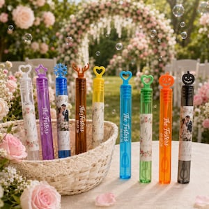 May include: A collection of eight cylindrical bubble wands in various colors, each topped with a unique shape like a heart, star, snowflake, deer antlers, or pumpkin. The wands feature personalized labels with floral designs, the text The Fosters, A & J, and the date 08.07.26.